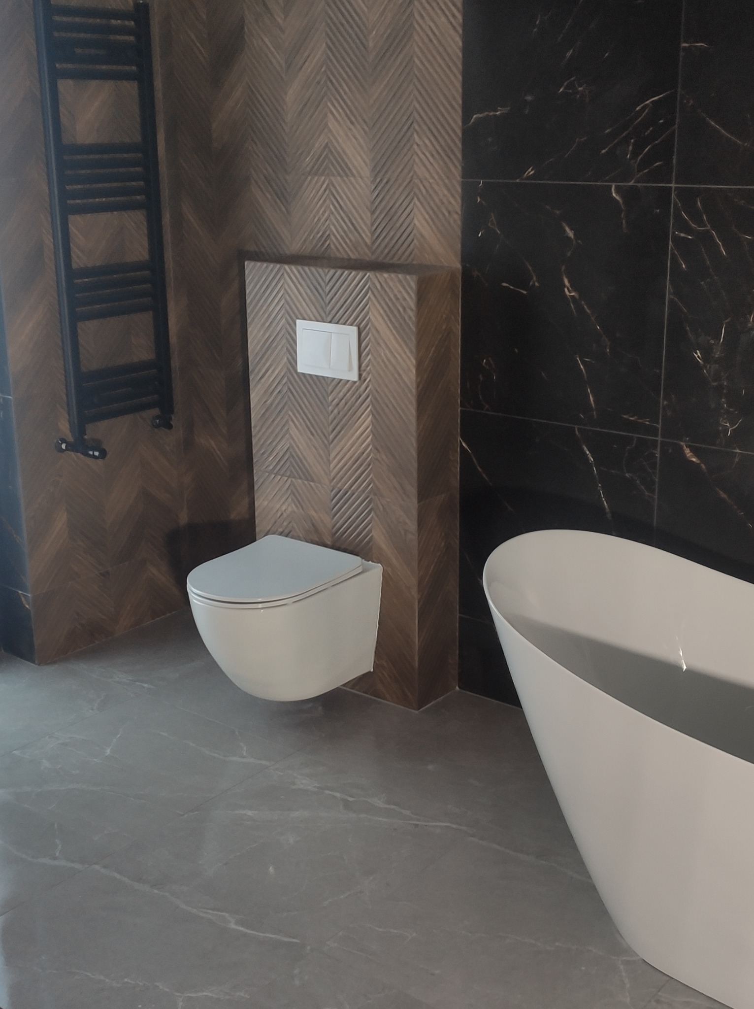 Elegancka łazienka z mozaiką w jodełkę na ścianie, podwieszana miska WC i fragment białej wanny na tle ciemnych płytek imitujących marmur.