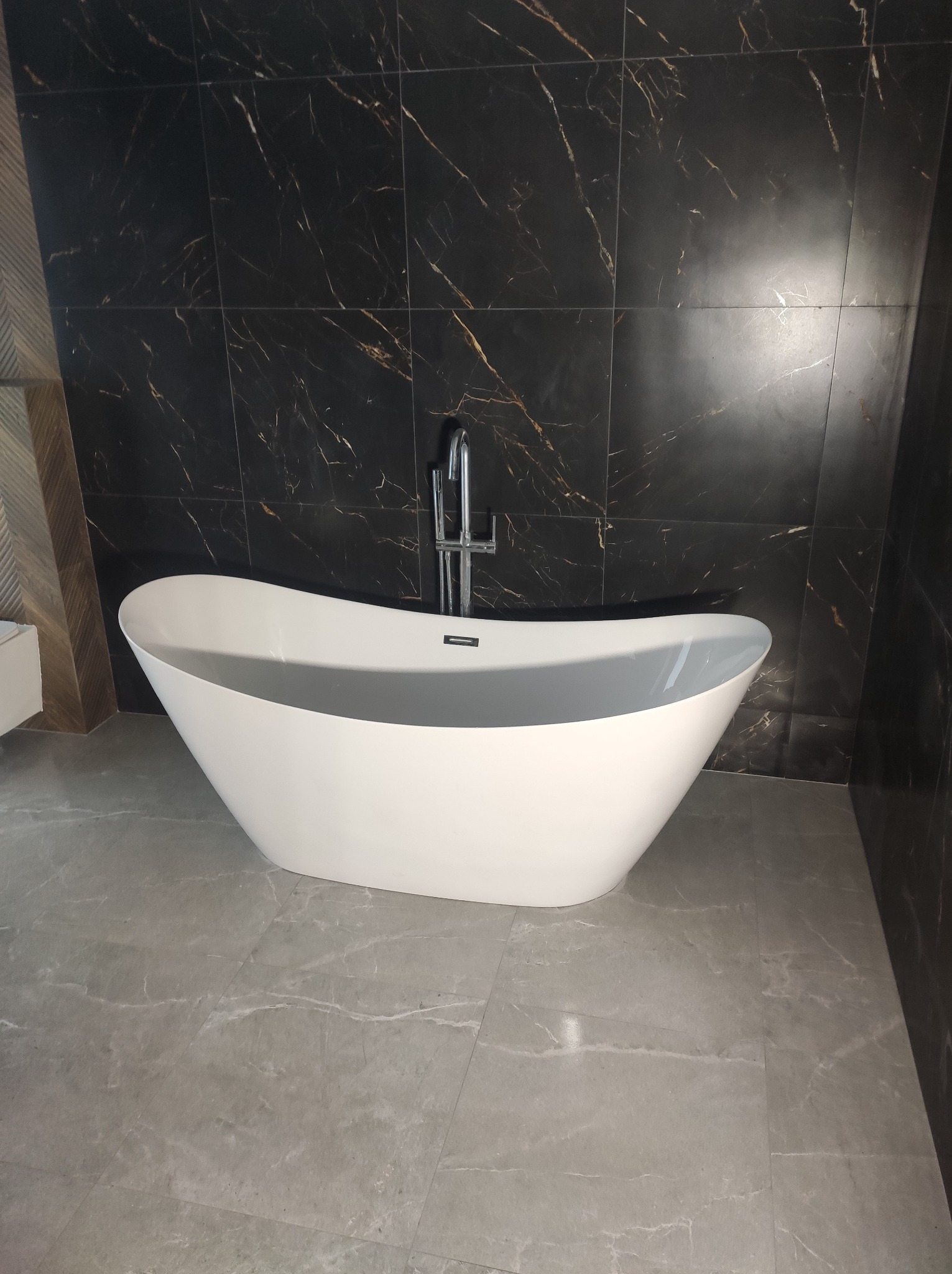 Biała wolnostojąca wanna wypełniona wodą na tle ściany z czarnego marmuru z złotymi żyłami i szarej marmurowej podłogi. Chromowana bateria.
