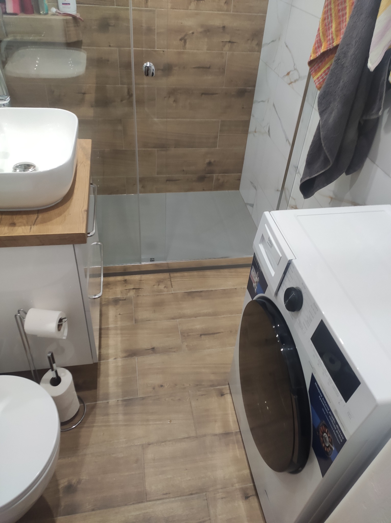 Nowoczesna łazienka po remoncie: drewnopodobne płytki, kabina prysznicowa, umywalka nablatowa i pralka. Widok na wykończone wnętrze z bliska.