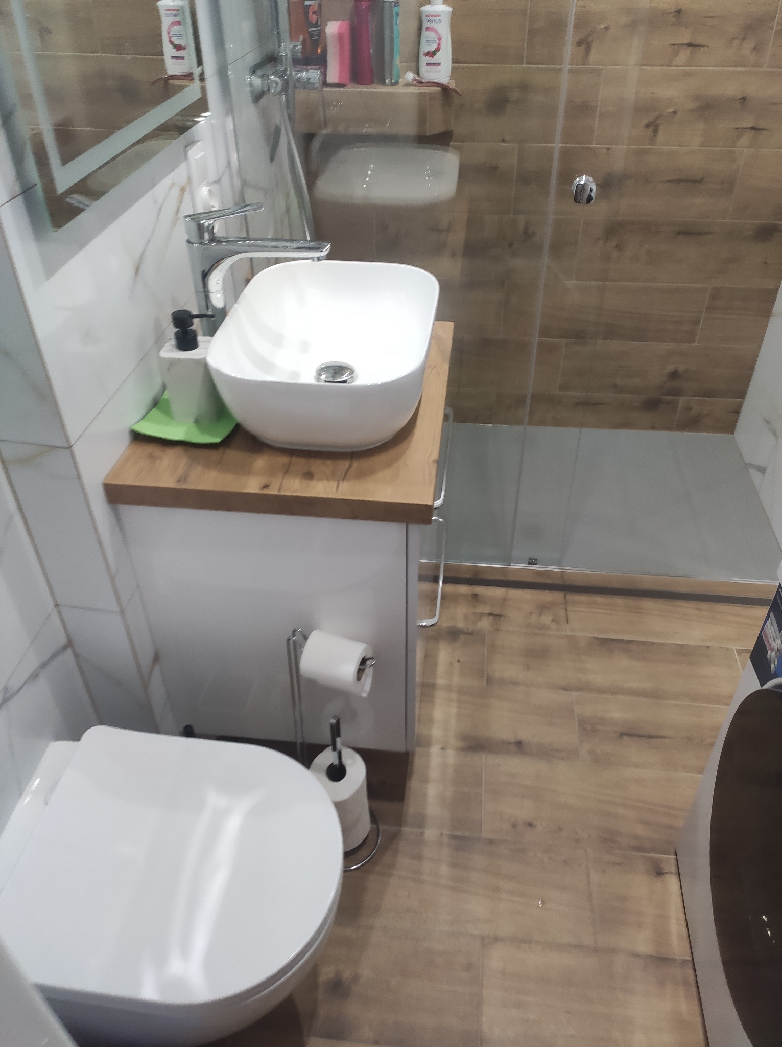 Nowoczesna łazienka z umywalką nablatową, drewnianym blatem, kabiną prysznicową i toaletą. Uwagę przykuwają płytki imitujące drewno na podłodze i ścianie.