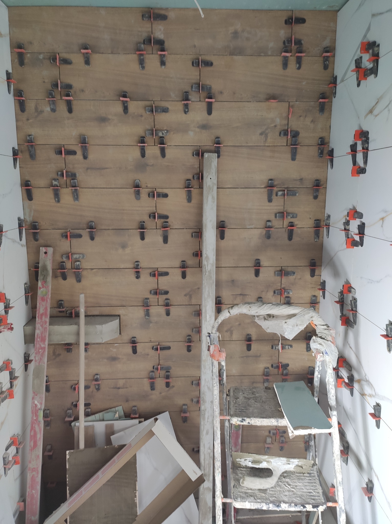 Ściana z drewnopodobnych płytek, w trakcie montażu z użyciem klipsów poziomujących. Widoczne narzędzia i drabina. Płytki ułożone poziomo.