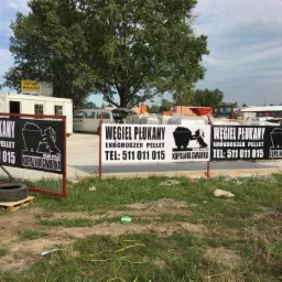 Reklama składu opału z banerami informującymi o sprzedaży węgla płukanego, ekogroszku i pelletu, umieszczona na ogrodzeniu z siatki, z widocznym magazynem i placem składowym w tle.