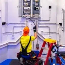 Elektryk w żółtej koszulce i pomarańczowej czapce, pracujący przy instalacji elektrycznej w białym pomieszczeniu, z widoczną skrzynką rozdzielczą i przewodami ułożonymi w listwach, drabina...