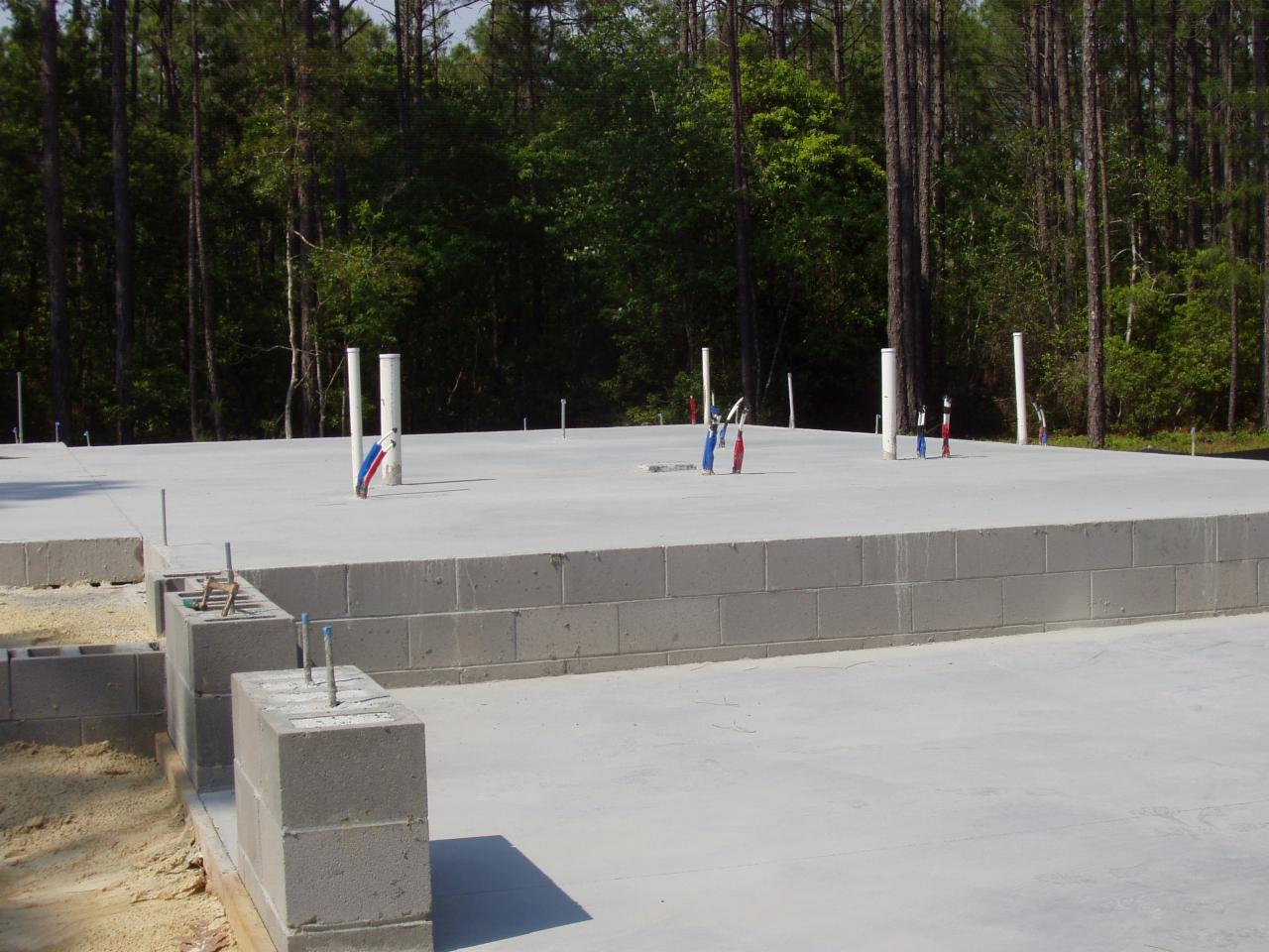 Betonowa płyta fundamentowa z wystającymi rurami instalacyjnymi i częściowo wzniesionymi ścianami z bloczków betonowych, teren zalesiony w tle.