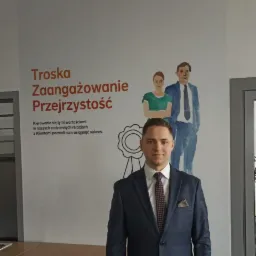 Mężczyzna w garniturze stoi na tle ściany z napisem 'Troska, Zaangażowanie, Przejrzystość' i ilustracją rodziny.