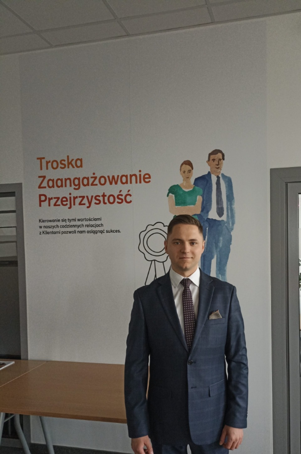 Mężczyzna w garniturze stoi na tle ściany z napisem 'Troska, Zaangażowanie, Przejrzystość' i ilustracją rodziny.