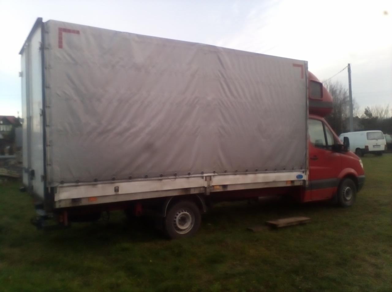 Czerwony samochód dostawczy Sprinter 318CDI z plandeką na trawiastym terenie. Widoczny fragment skrzyni ładunkowej z szarą plandeką, elementy zawieszenia i opona. W tle budynki i inne pojazdy.