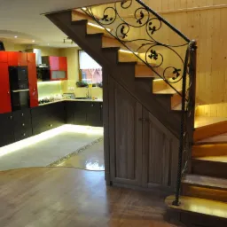 Nowoczesna kuchnia z czerwono-czarnymi szafkami, drewnianymi schodami z dekoracyjną metalową balustradą i podświetleniem LED pod szafkami oraz stopniami.