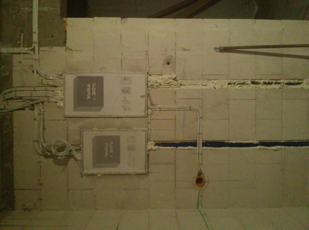 Dwa panele elektryczne 'Volta +30%' zamontowane na ścianie z widocznymi kablami i przewodami w trakcie instalacji, z pianką montażową w szczelinach.