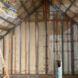 Poddasze w trakcie izolacji pianą, widoczne metalowe profile konstrukcyjne, drewniane elementy więźby dachowej i wisząca lampa budowlana.