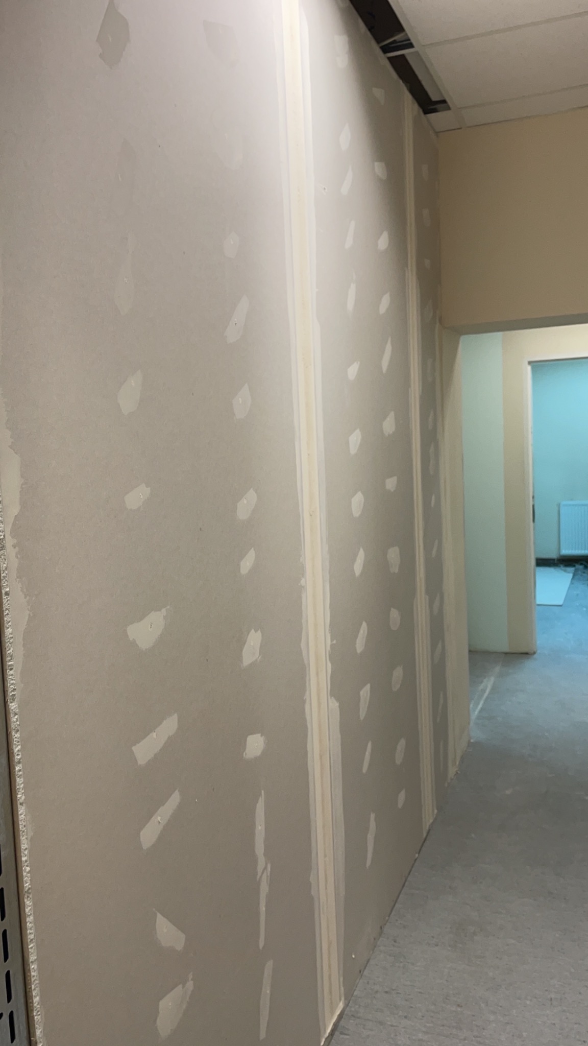 Ściana z płyt gipsowo-kartonowych w trakcie szpachlowania, widoczne miejsca po wkrętach wypełnione masą, perspektywa z dołu, w tle widoczne otwarte przejście do innego pomieszczenia.