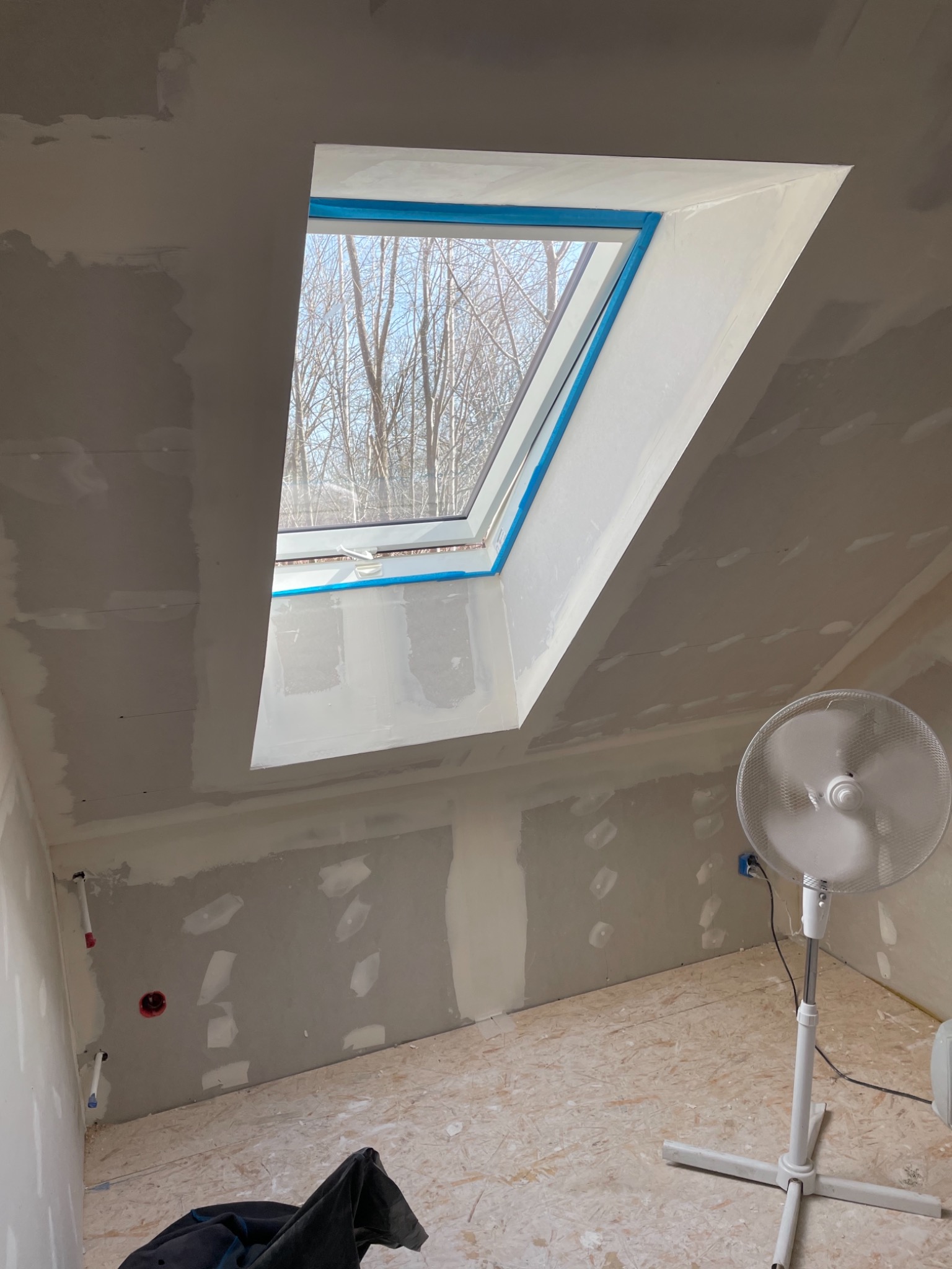 Pokój na poddaszu w trakcie wykańczania: ściany i sufit częściowo pokryte masą szpachlową, widoczne łączenia płyt, okno dachowe zabezpieczone niebieską taśmą, na podłodze leżą ubrania robocze, a...