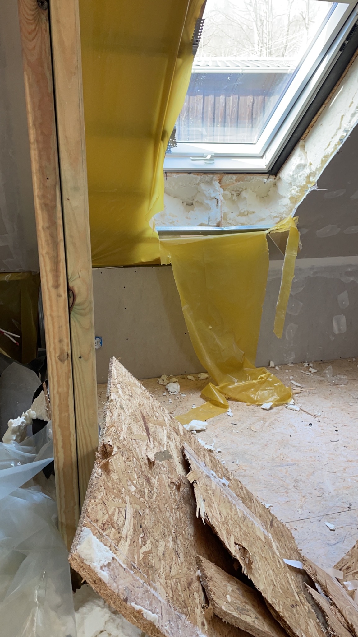 Montaż okna dachowego: widok na surowy otwór okienny z żółtą folią ochronną, fragmentem ściany pokrytej płytą, drewnianą ramą i leżącymi na podłodze fragmentami płyty OSB z resztkami pianki...