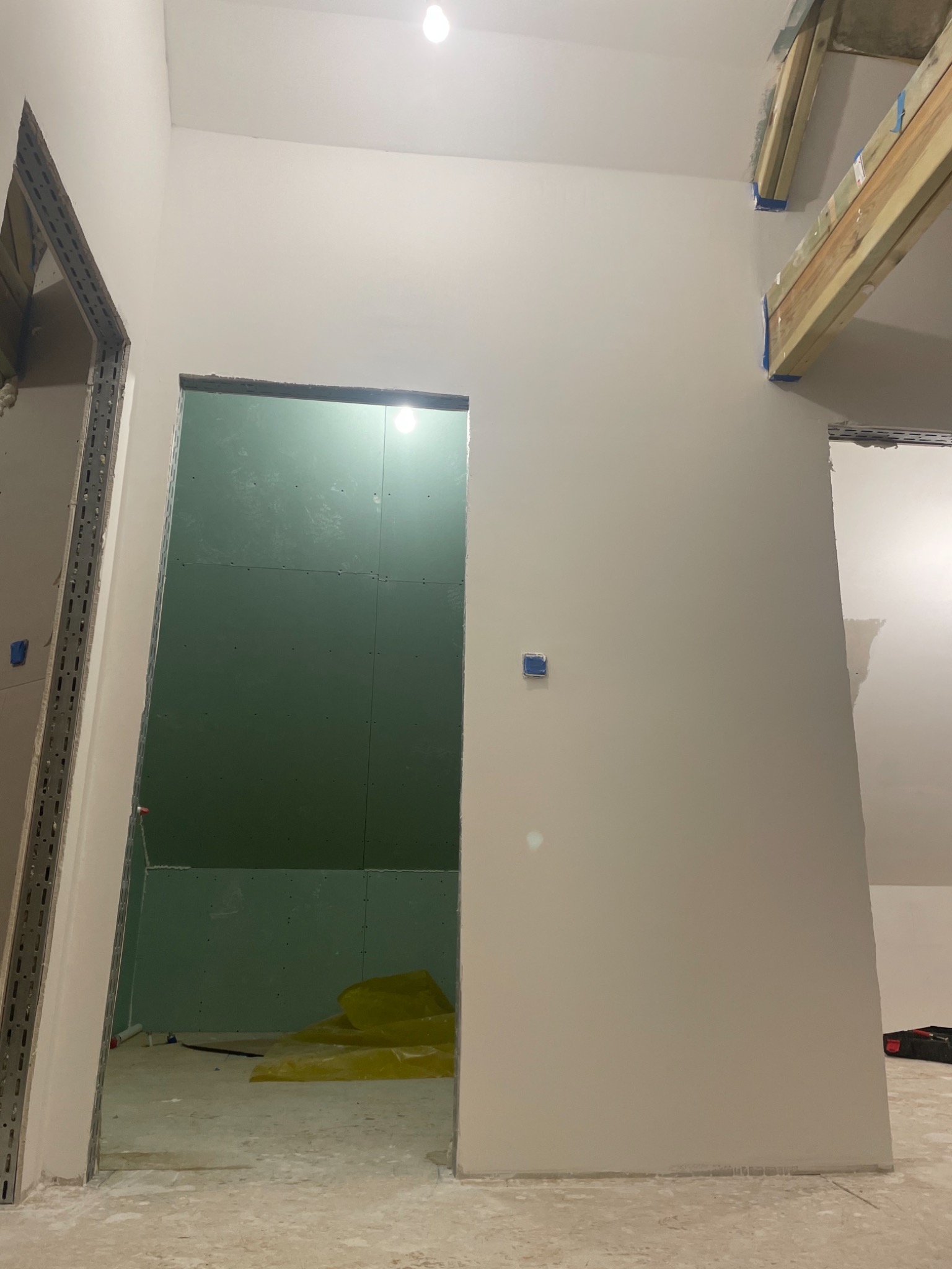 Wnętrze poddasza w trakcie wykańczania: widoczne stelaże metalowe przy otworze drzwiowym, ściany częściowo pokryte zielonymi płytami, zabezpieczona folią podłoga, drewniane elementy konstrukcyjne...