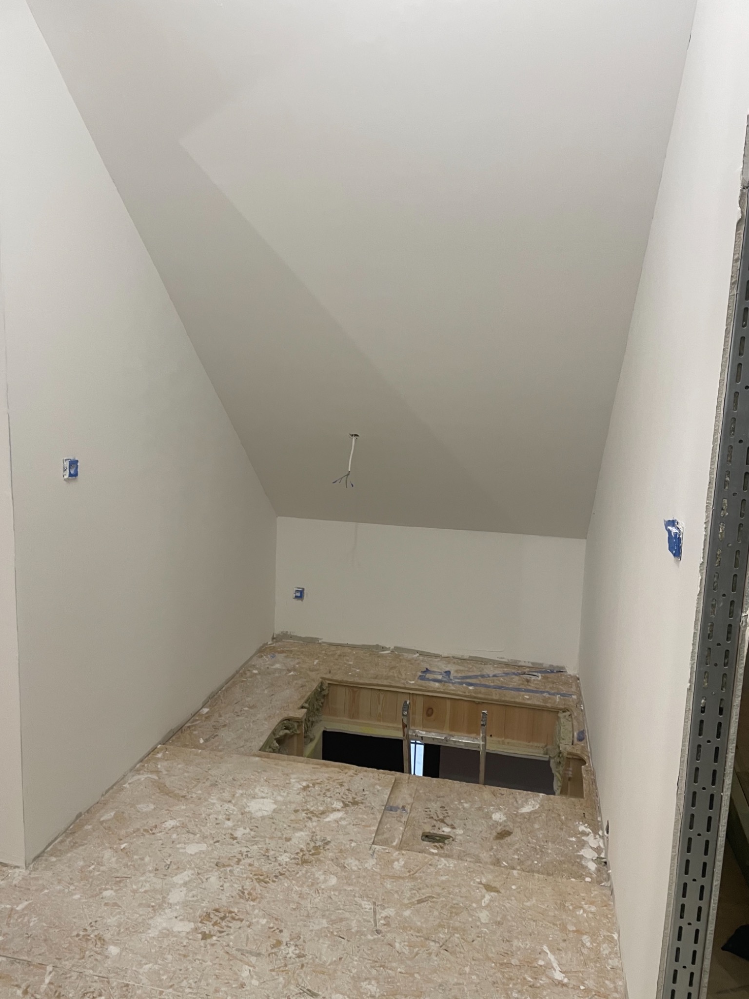 Wnętrze poddasza w trakcie remontu z odsłoniętą konstrukcją podłogi i świeżo pomalowanymi ścianami na biało, widoczne puszki elektryczne i wystające przewody.