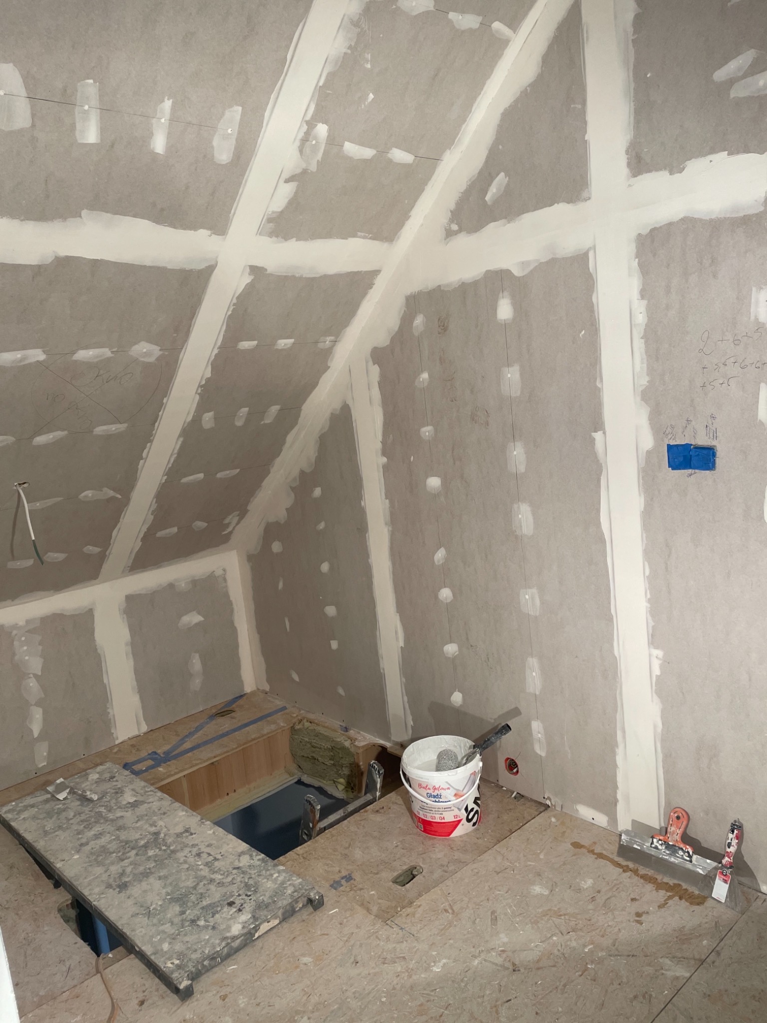 Poddasze w trakcie wykańczania: płyty karton-gips na skośnych ścianach i suficie, widoczne łączenia wzmocnione taśmą i wstępnie wypełnione masą szpachlową, na podłodze wiadro z gładzią i szpachelki.