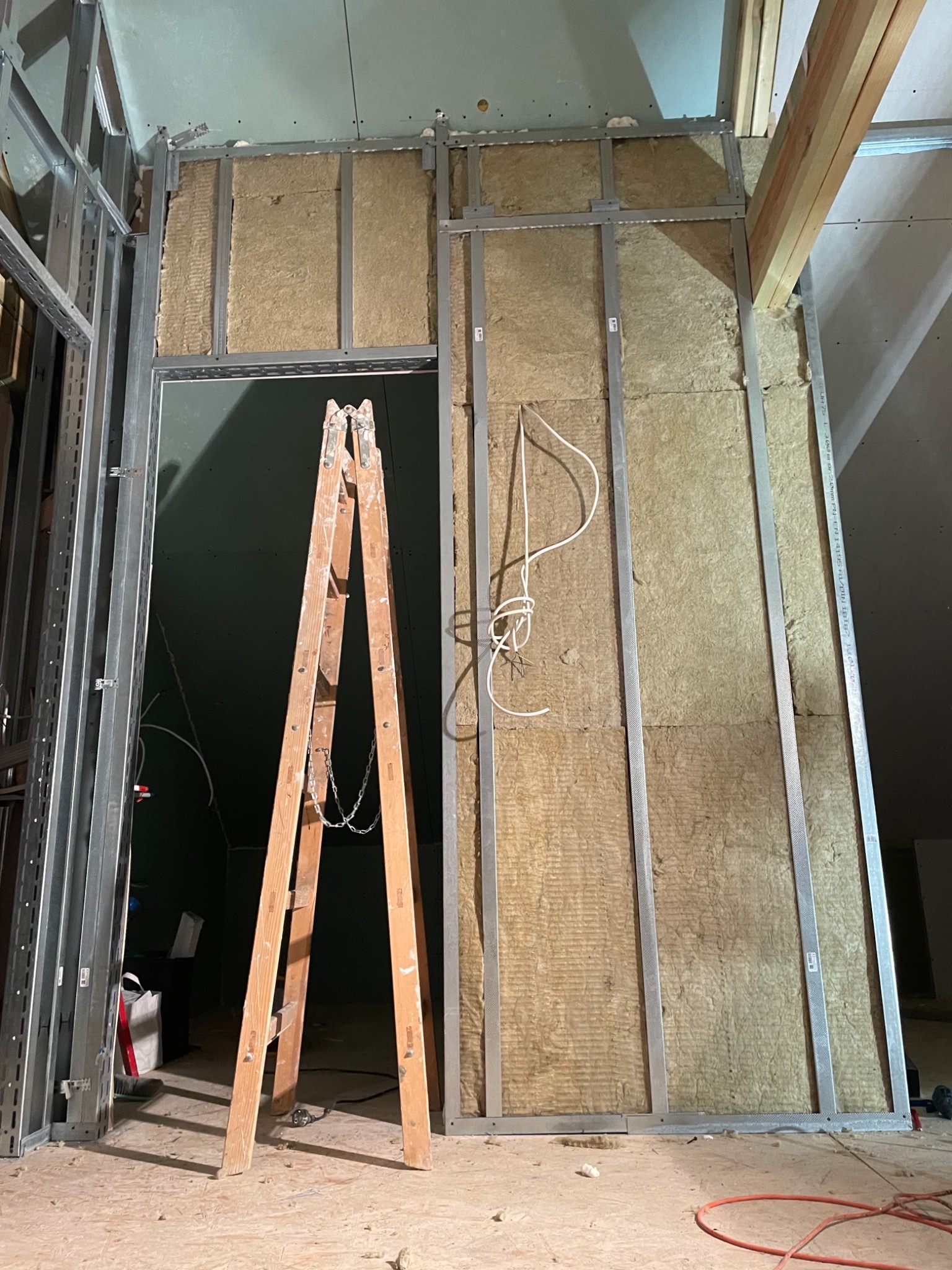 Montaż izolacji z wełny mineralnej w metalowym stelażu ściany działowej, widoczna drabina malarska i przewody elektryczne w surowym wnętrzu poddasza.