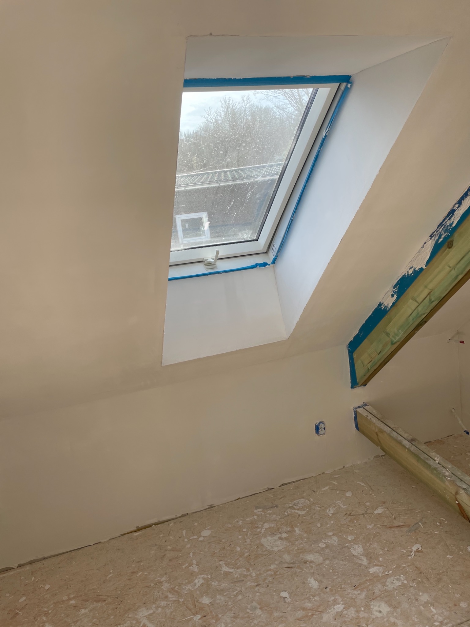 Poddasze z oknem dachowym po świeżo nałożonej gładzi, zabezpieczone taśmą malarską, widoczne ślady gładzi na podłodze.