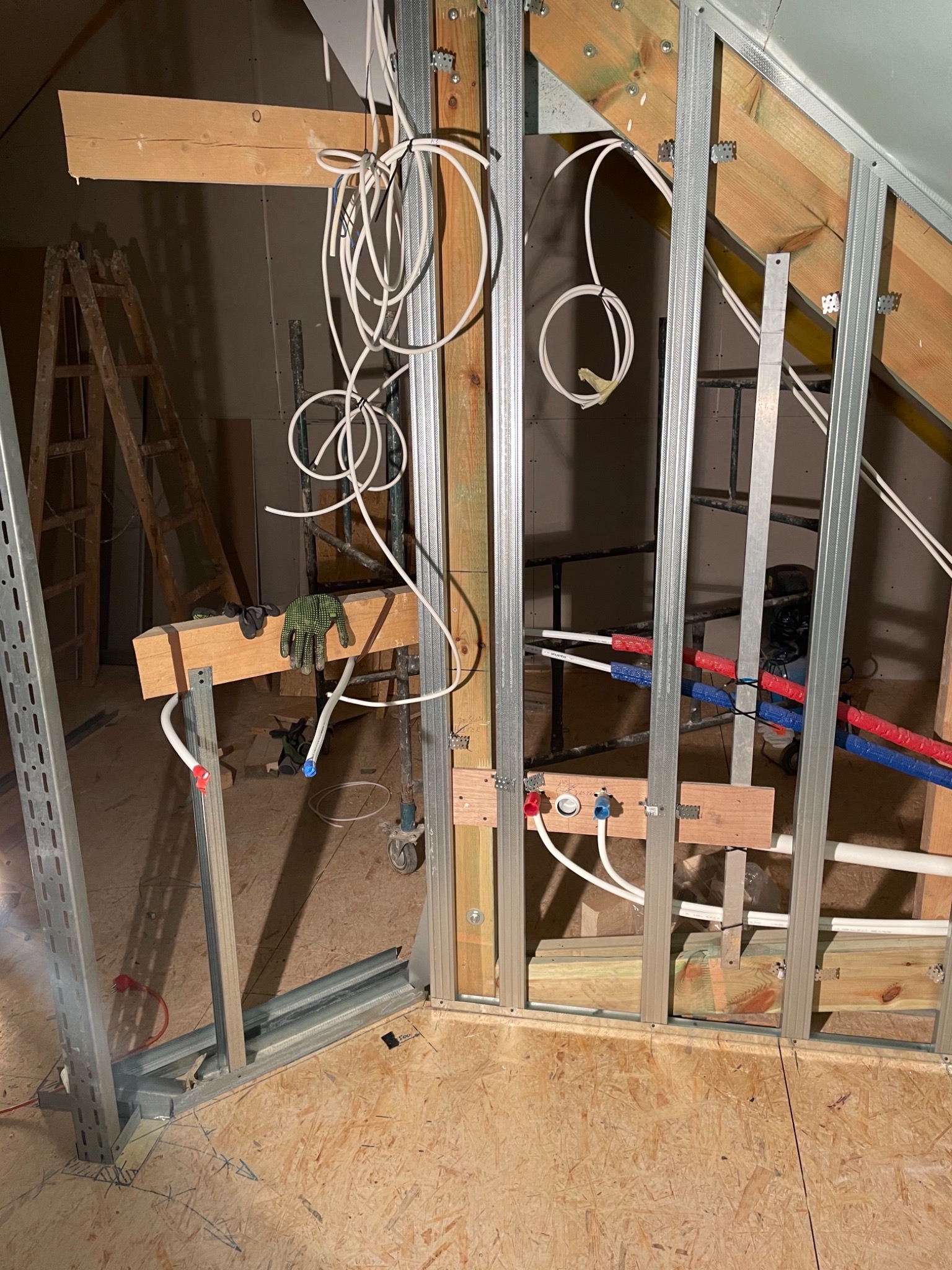 Konstrukcja szkieletowa z metalowych profili, drewnianych elementów i instalacji wodnej w trakcie budowy poddasza; widoczne przewody elektryczne, rury wodne z izolacją, drabina i rękawice robocze.