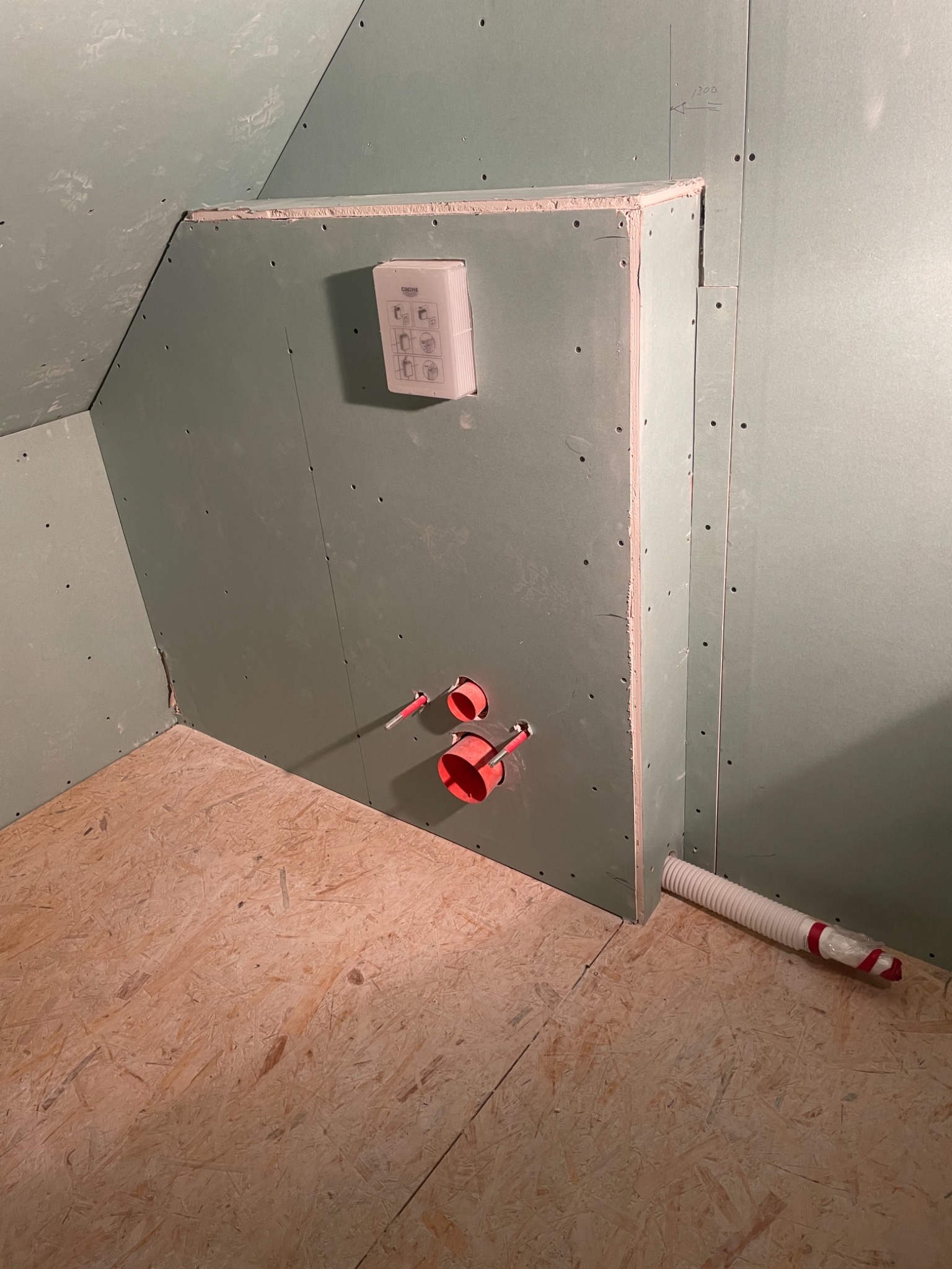 Wnętrze poddasza z ukośną ścianą, wykończone surowymi płytami gipsowo-kartonowymi, widoczne otwory na instalacje sanitarne i elektryczne, podłoga z płyt OSB, w rogu biała rura karbowana.