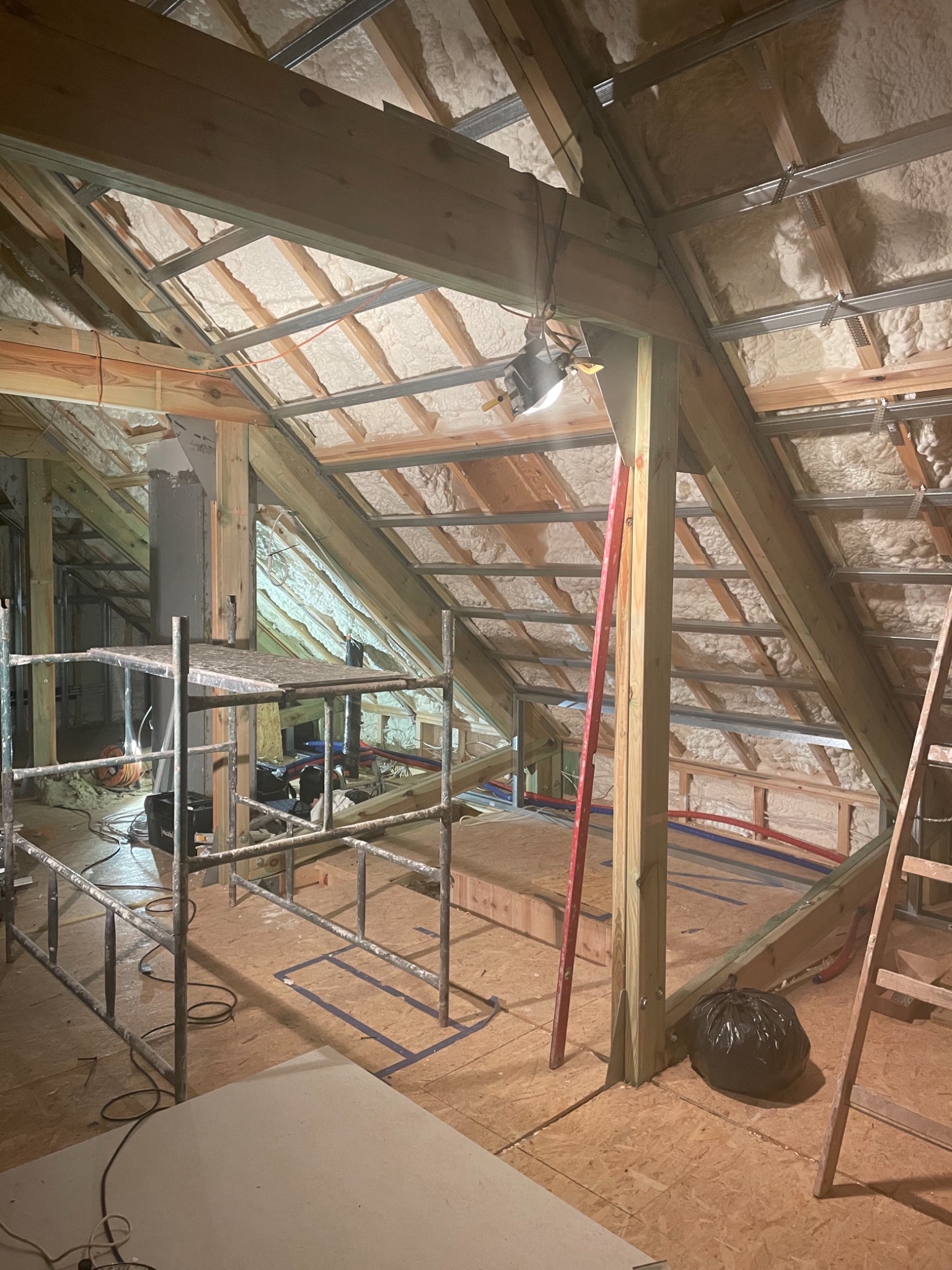 Poddasze w trakcie adaptacji: ocieplenie wełną mineralną, drewniana konstrukcja dachu, metalowe profile, instalacje, rusztowanie, płyta budowlana na podłodze i lampa robocza.