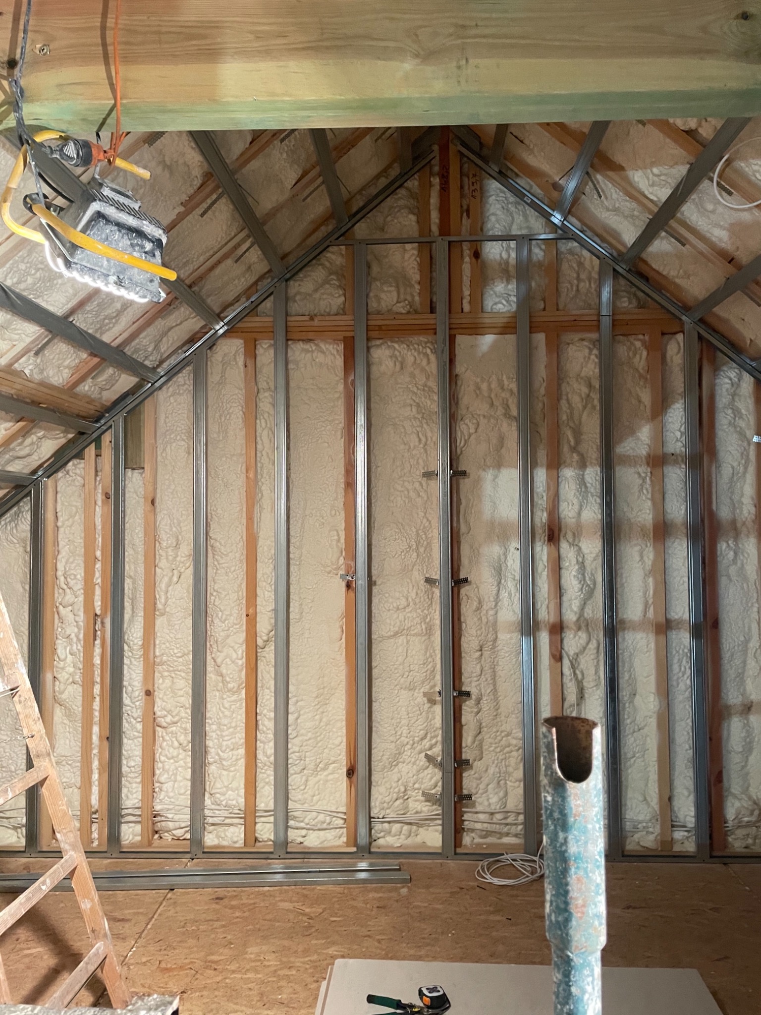 Poddasze w trakcie izolacji pianą, widoczne metalowe profile konstrukcyjne, drewniane elementy więźby dachowej i wisząca lampa budowlana.