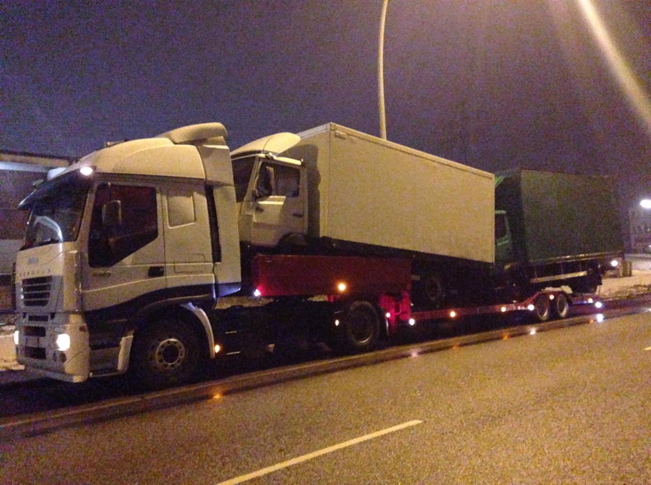 Laweta przewozi dwa samochody ciężarowe w nocy, widoczne światła ostrzegawcze i oświetlenie uliczne.