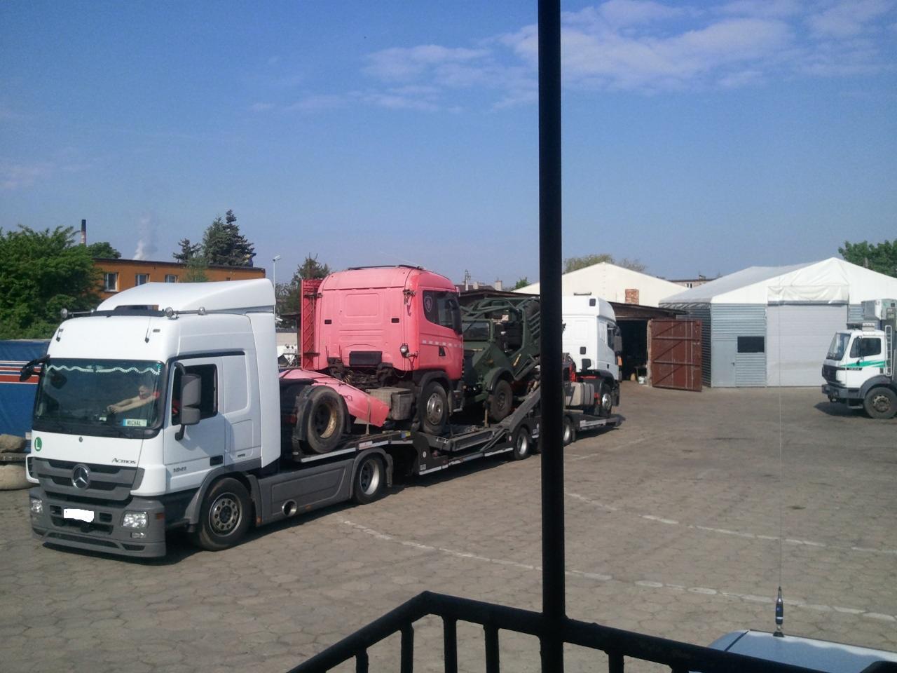 Ciężarówka laweta przewozi inne ciężarówki na parkingu przemysłowym w słoneczny dzień.