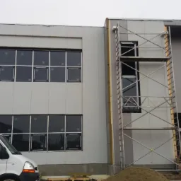 Budynek w trakcie budowy z szarych płyt warstwowych, widoczne rusztowanie i biały samochód dostawczy.