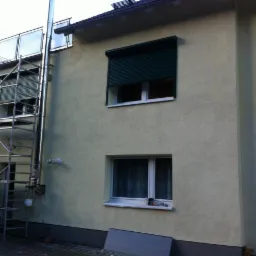Elewacja budynku z dwoma oknami, jedno z zieloną roletą częściowo opuszczoną, drugie z firanką, widoczne rusztowanie i rura kominowa na zewnątrz, przy elewacji leży szara płyta.