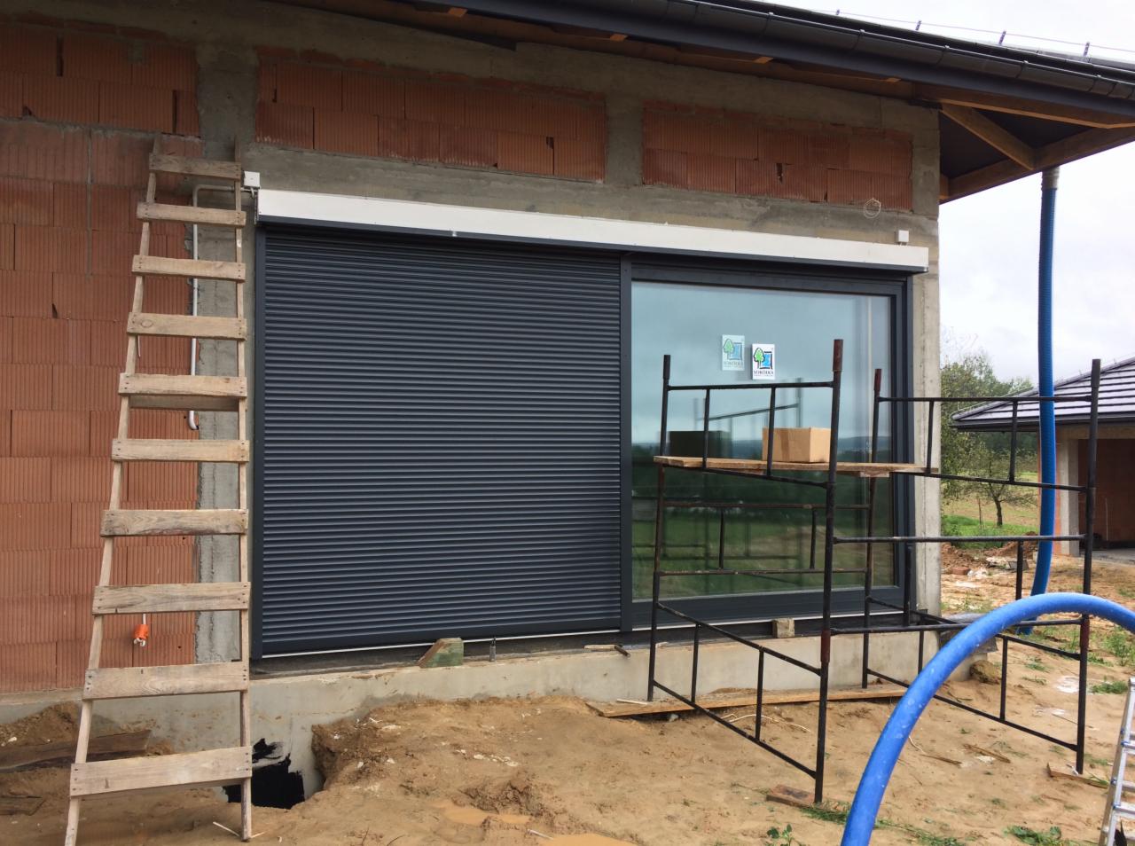 Okno z roletą zewnętrzną w trakcie montażu w surowym stanie budynku, widoczne rusztowanie, drabina oparta o ścianę z cegły dziurawki i niezagospodarowany teren wokół.