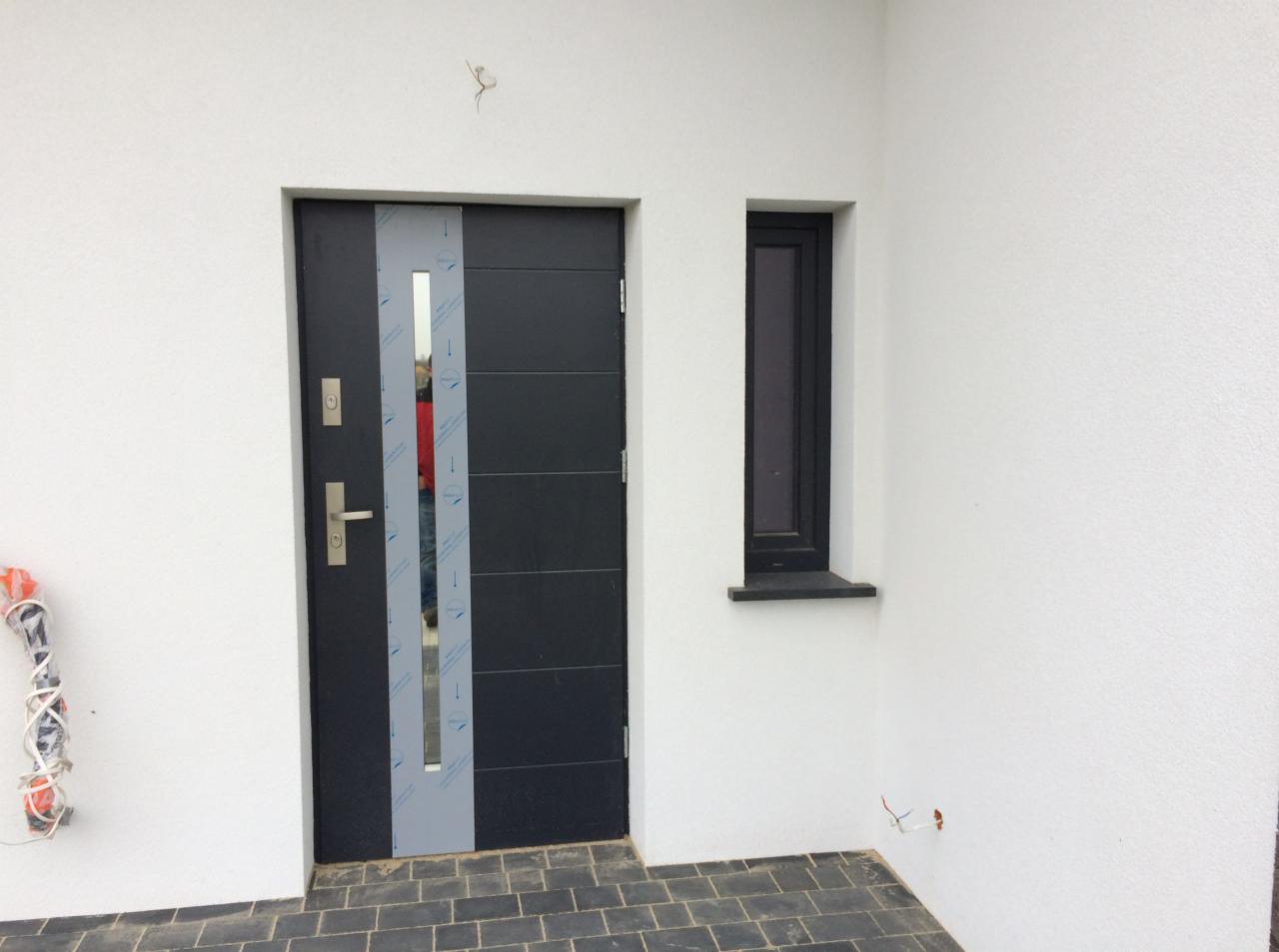 Nowoczesne, ciemnoszare drzwi wejściowe z metalowymi akcentami i wąskim oknem, częściowo zabezpieczone folią ochronną, zamontowane w białej ścianie obok pionowego okna, widoczne przewody...