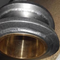 Camet-system - Metalowy element z widocznymi śladami spawania i mosiężną tuleją wewnątrz, leżący na drewnianej powierzchni.