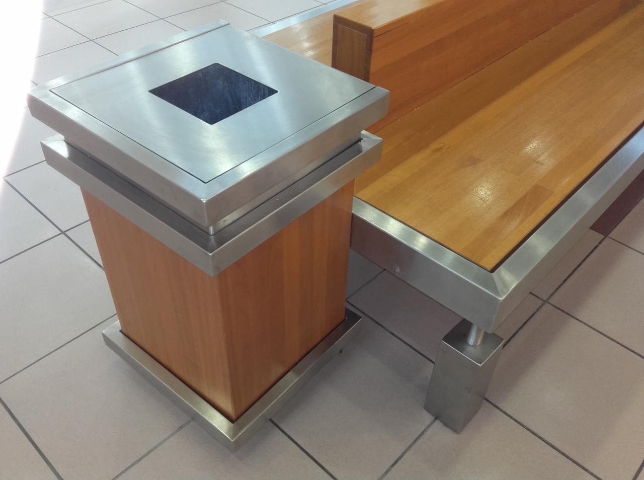 Stylowy kosz na śmieci z drewnianą obudową i metalowym wykończeniem, stojący obok ławki z drewnianym siedziskiem i metalową ramą, na tle jasnej podłogi z płytek.