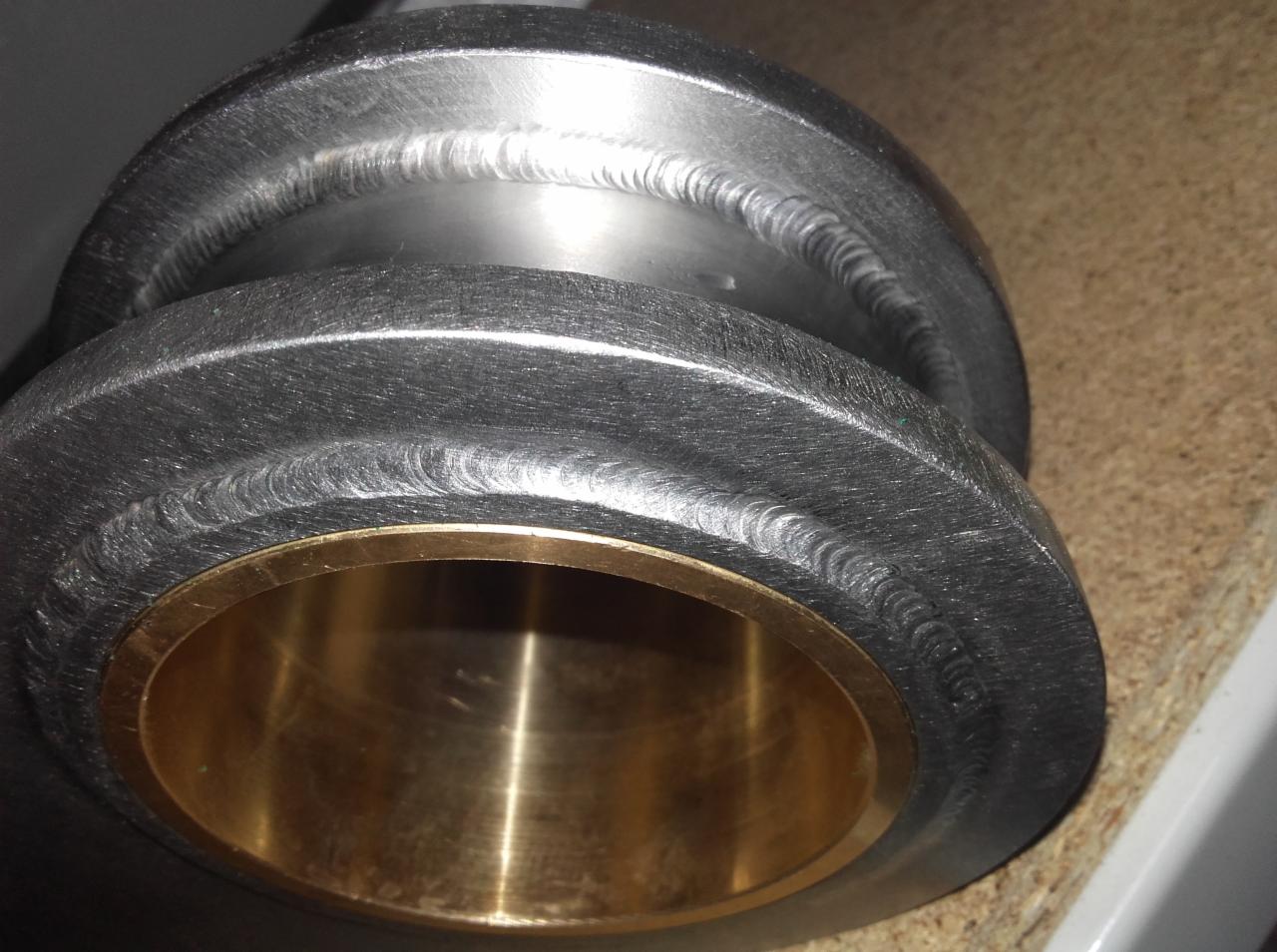 Metalowy element z widocznymi śladami spawania i mosiężną tuleją wewnątrz, leżący na drewnianej powierzchni.