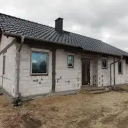 Surowy dom jednorodzinny w trakcie budowy z widocznymi oknami PCV z białymi ramami, czarnym dachem i rynnami, na tle pochmurnego nieba i niezagospodarowanego terenu.