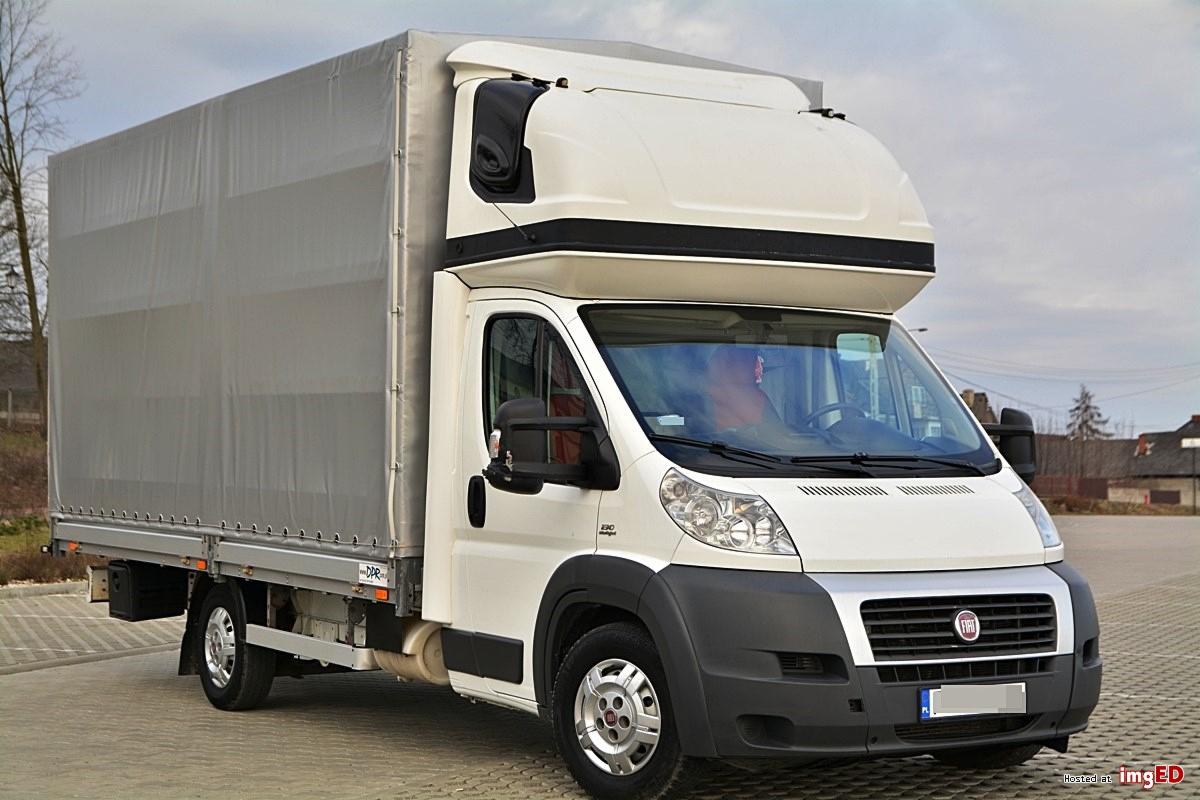 Biały samochód dostawczy Fiat Ducato z plandeką na parkingu, widok z przodu, z widocznymi elementami mocowania plandeki.