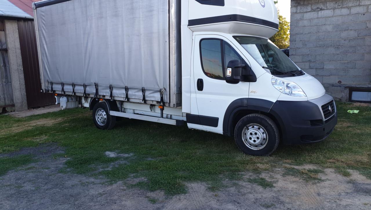 Biały samochód dostawczy z plandeką na trawniku, widok z boku. Lekko pochylony kadr, szara plandeka i zabudowa nad kabiną.