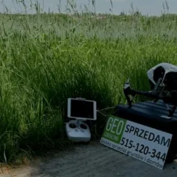 Dron DJI Inspire 2 na czarnym kufrze obok tabliczki z napisem 'GEO Consulting Sprzedam 515-120-344 Szybka sprzedaż grunt&oacute;w i dom&oacute;w', obok leży tablet z kontrolerem, w tle wysoka trawa.