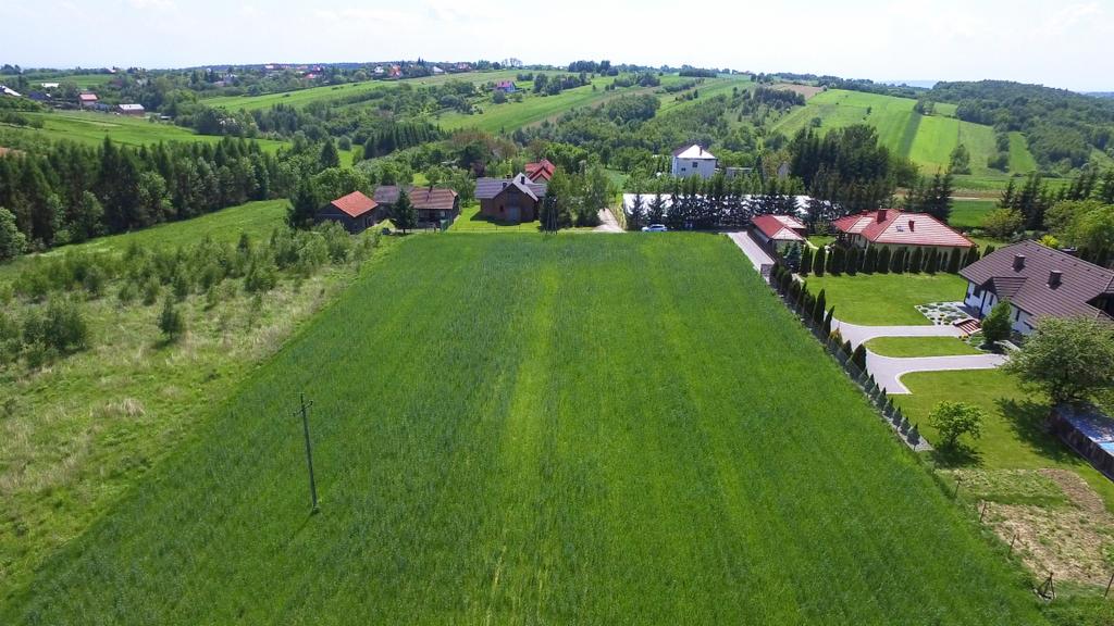 Panoramiczny widok z lotu ptaka na działkę budowlaną z zielonym polem uprawnym w pierwszym planie, w tle domy jednorodzinne i zalesione wzgórza.