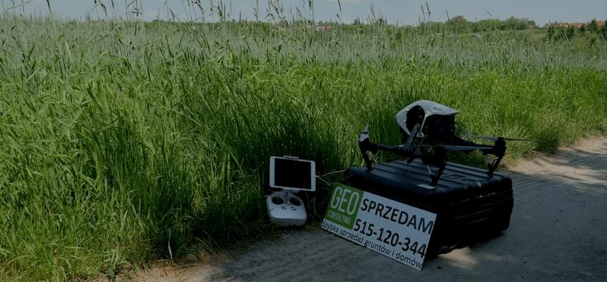 Dron DJI Inspire 2 na czarnym kufrze obok tabliczki z napisem 'GEO Consulting Sprzedam 515-120-344 Szybka sprzedaż gruntów i domów', obok leży tablet z kontrolerem, w tle wysoka trawa.