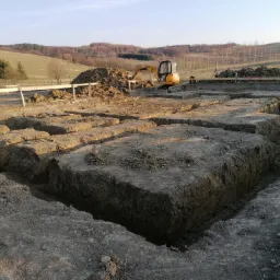 Żółta mini koparka JCB przy wykopach pod fundamenty w Sanoku, na tle pagórkowatego krajobrazu i lasu.