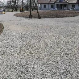 Szeroki kadr na utwardzony żwirem teren posesji z widocznym domem w tle, zagęszczarką w oddali i fragmentem ogrodzenia po lewej stronie.