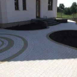 Ułożona kostka brukowa przed domem z elementami dekoracyjnymi w kształcie okręgów i półokręgów, obrzeża z kostki w innym odcieniu, przygotowane rabaty ziemi.