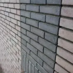 Fragment ściany z cegły klinkierowej w trzech odcieniach: jasny beż, szary i brązowy. Widoczne różnice w kolorystyce i fakturze poszczególnych partii muru.