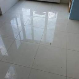 JLInwesting - Połyskująca, jasna podłoga wyłożona kwadratowymi płytkami ceramicznymi, odbijająca światło okna, z widocznymi fugami i drobnymi zabrudzeniami.