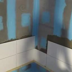 JLInwesting - Fragment łazienki w trakcie układania białych, prostokątnych płytek na ścianach, widoczne ślady kleju i przygotowane podłoże z niebieską izolacją przeciwwilgociową.