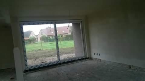 Surowy stan wnętrza domu w trakcie budowy z dużym oknem przesuwnym z widokiem na ogród i sąsiednie domy, ściany w szarym tynku, betonowa podłoga.