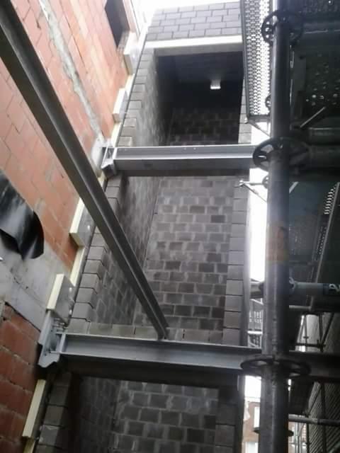 Wznoszenie muru z szarych bloczków betonowych z widocznymi stalowymi belkami wzmacniającymi konstrukcję, ujęcie z dołu z fragmentem rusztowania po prawej stronie.