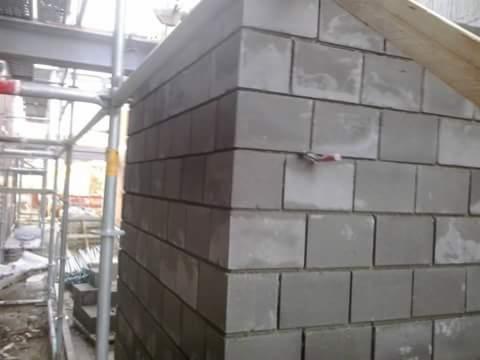 Wznoszenie ściany z szarych bloczków betonowych, widoczne rusztowanie i drewniany element konstrukcyjny, zaprawa pomiędzy bloczkami jeszcze świeża.